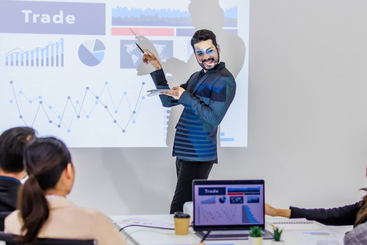 Millennial Asian young cheerful male businessman presenter in formal suit standing holding notebook and pen pointing presenting corporate graph chart data information from wall screen in meeting room