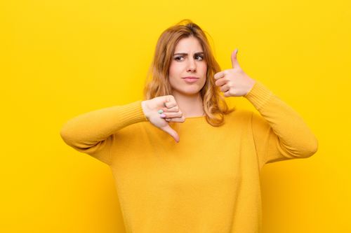 young pretty blonde woman feeling confused, clueless and unsure, weighting the good and bad in different options or choices against yellow wall